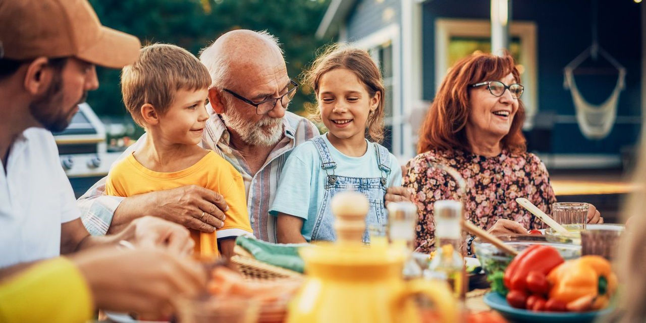Portrait of a Happy Senior Grandfather Holding His Bright Talented Little Grandchildren on Lap at a Outdoors Dinner Party with Food and Drinks. Family Having a Picnic Together with Children.