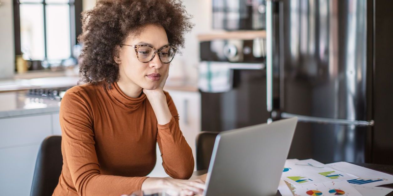 Young woman seated at kitchen table looking at her investments on a laptop