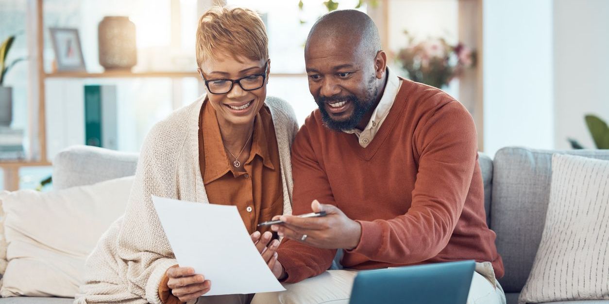 Finance, paperwork and happy black couple on sofa check bank account, financial statements and report. Budget, saving and middle aged man and woman do online banking on laptop with documents at home