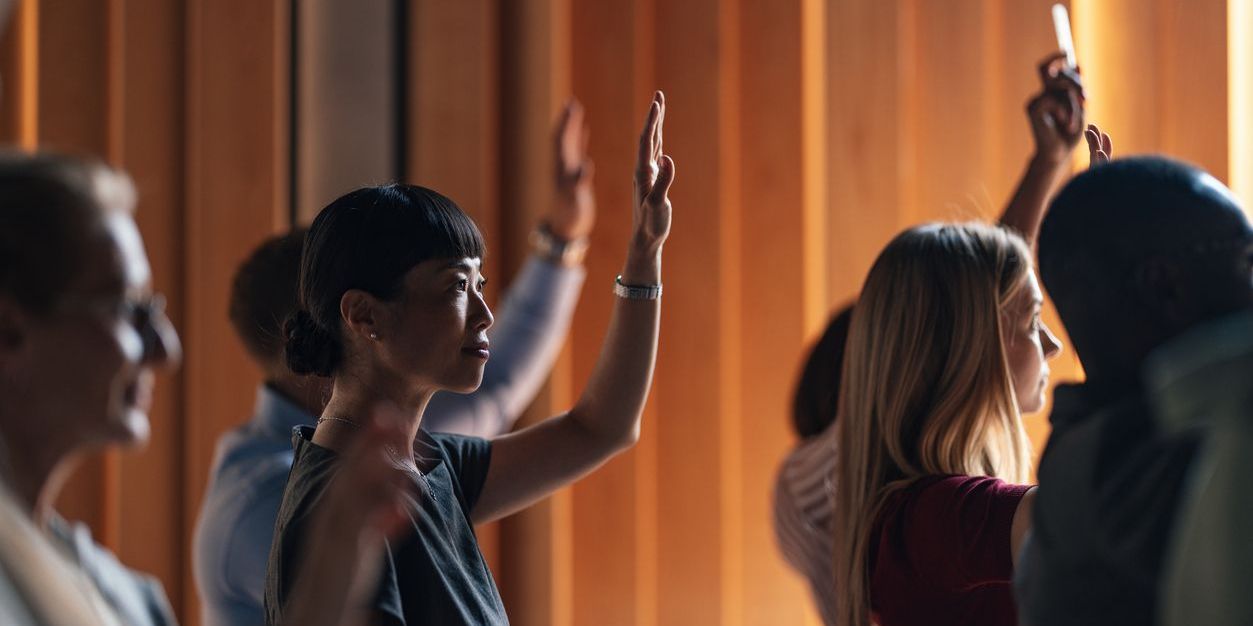 A diverse group of individuals actively participating in a business lecture. Several attendees are raising their hands, demonstrating engagement and interest in the discussion.
