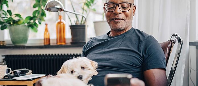 Middle-aged Black man at home relaxing in a chair and looking at this phone while his small white dog rests in his lap.t his phone.