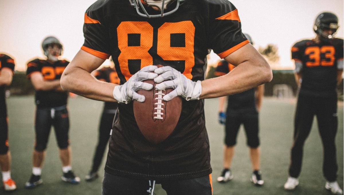Close-up photo of a football player holding a football with teammates in the background
